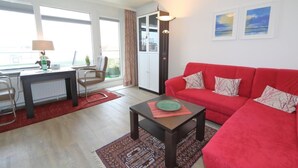 A living room with a red sofa, a small table, a lamp, and a view of the outdoors through large windows. - Haus am Meer14-app 078a OB (Sylt)