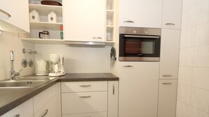 A compact kitchen with white cabinets, a stainless steel oven, a coffee maker, and a sink. - Haus am Meer14-app 078a OB (Sylt)