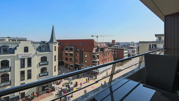 A view from a balcony overlooking a city street with buildings, including a 'MENSING' sign, and a construction crane in the distance. - Haus am Meer14-app 078a OB (Sylt)
