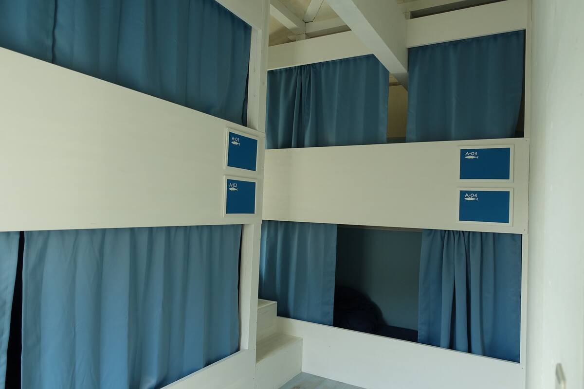 Ein Schlafraum mit Etagenbetten, blauen Vorhängen und beschrifteten Fächern A-01, A-02, A-03 und A-04.