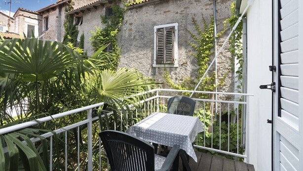 Un balcone con tavolo e sedie, immerso nel verde e affacciato su un edificio in pietra con tetto in tegole.