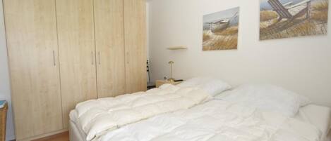 A bedroom with a large wardrobe, a bed with white bedding, and two landscape paintings on the wall.