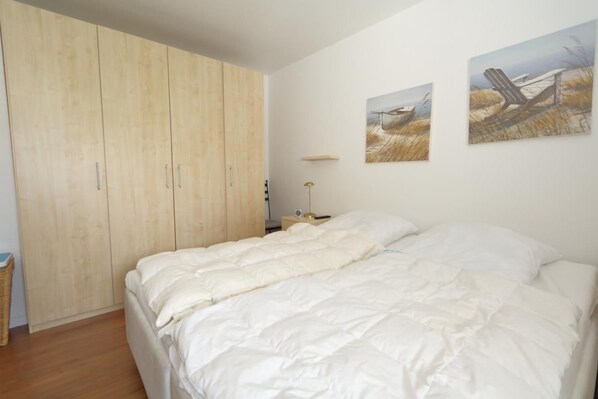 Une chambre avec une grande armoire, un lit avec une literie blanche et deux tableaux représentant des paysages accrochés au mur.
