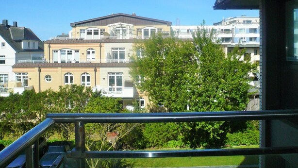 En bostadsbyggnad med en balkong, omgiven av grönska och andra byggnader.