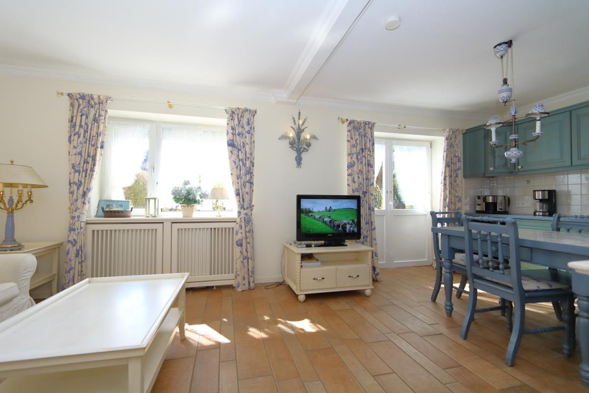 A well-lit living room with a television, a dining table with chairs, and a kitchen area with teal cabinets.