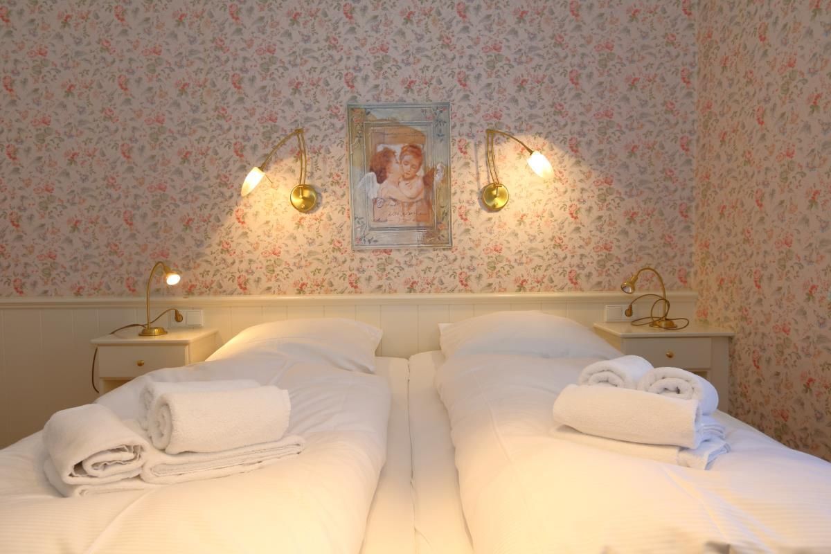 Een tweepersoonskamer met twee bedden, elk voorzien van keurig opgevouwen witte handdoeken, een ingelijst schilderij boven de bedden en twee bedlampen.