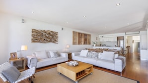 A spacious living room with a sofa set, a coffee table, and a dining area with a kitchen in the background. - The Quarter Deck - This riverfront townhouse is modern, fresh and spacious (Port Fairy)
