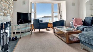 Een gezellige woonkamer met een stenen muur, een groot raam met uitzicht op de zee, een flatscreen televisie, twee blauwe fauteuils, een houten salontafel en een blauwe bank.