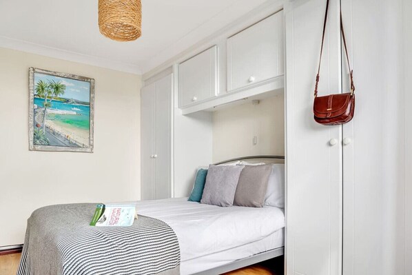 A bedroom with a bed, a white wardrobe, a hanging bag, and a beach-themed painting on the wall.