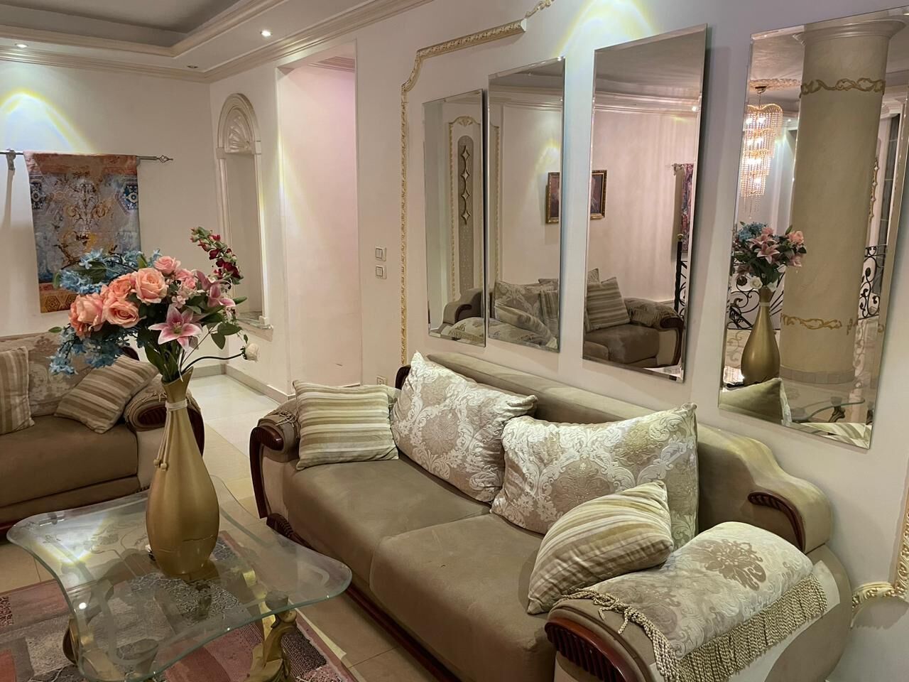 Una sala de estar con un sofá color beige, almohadones decorativos, una mesa de centro de vidrio y un jarrón con flores.