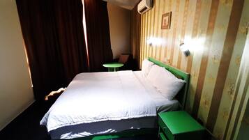 Quarto de hotel com cama, mesa de cabeceira verde, pequena mesa e luminária fixada na parede.
