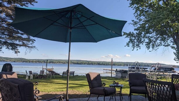 An outdoor patio with a view of a lake, featuring a large umbrella, wicker chairs, and a small table. - Beautiful lakefront cottage on Chautauqua Lake in a quiet setting! (Jamestown)