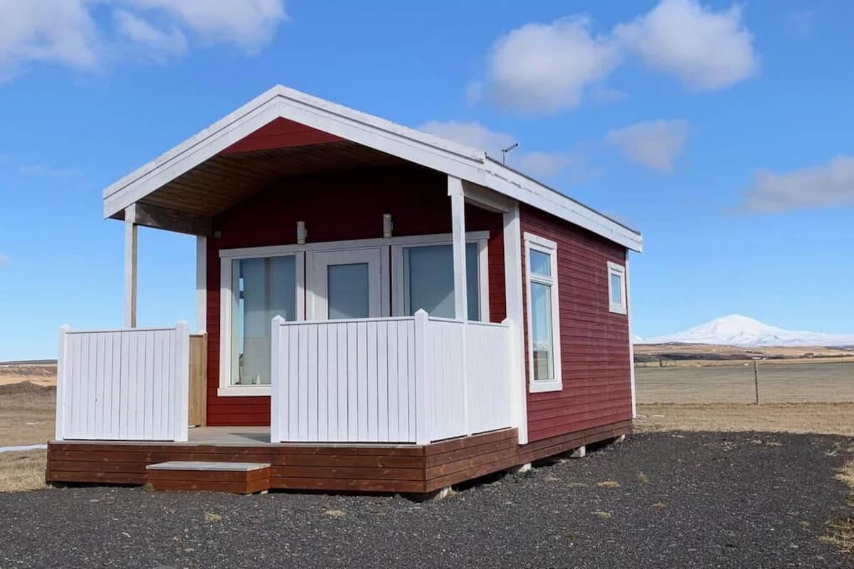 A small, single-story cabin with a red exterior and white trim, featuring a porch with white railings and a wooden deck.