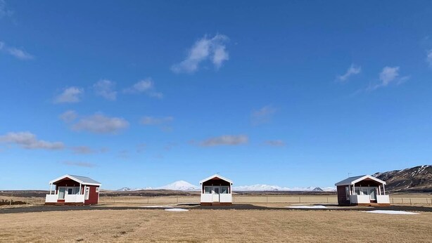 Zwei kleine, eingeschossige Holzhütten mit roter und weißer Fassade, eingebettet in eine flache, offene Landschaft mit Bergen im Hintergrund unter blauem Himmel.