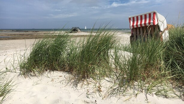 A beach scene with a beach chair covered in a red and white striped cloth, sand dunes, and a lighthouse in the distance.
