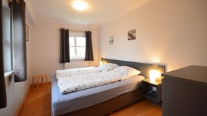 Un dormitorio con cama, mesita de noche, un taburete pequeño, ventana con cortinas y un radiador.