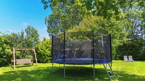 Un trampolín con red de seguridad, un columpio de madera y dos sillas de jardín en un patio cubierto de césped con árboles al fondo.