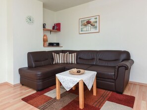 Ein Wohnzimmer mit braunem Ledersofa, einem kleinen Holztisch, einer Uhr, einer Lampe, einem gerahmten Bild und einem Bücherregal.