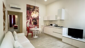 Una sala de estar moderna con un sofá blanco, una mesa de comedor con sillas, una zona de cocina con microondas y estufa, y un cartel grande en la pared.