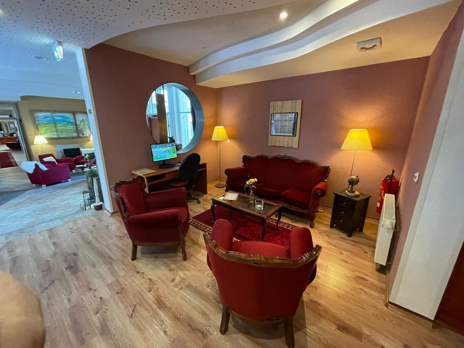 A cozy living room with a wooden floor, red armchairs, a red sofa, a round mirror, and a desk with a computer.