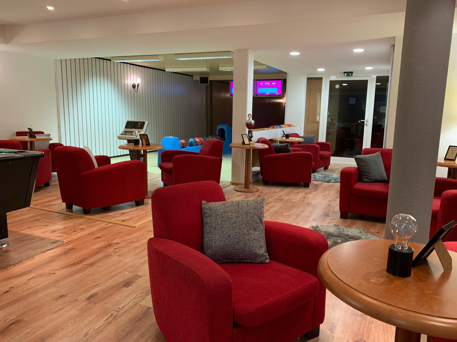 A modern lounge area with red upholstered chairs, a pool table, and a billiards table. There is a blue play area in the background and a television screen mounted on the wall.