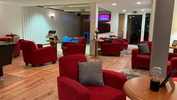 A modern lounge area with red upholstered chairs, a pool table, and a billiards table. There is a blue play area in the background and a television screen mounted on the wall.