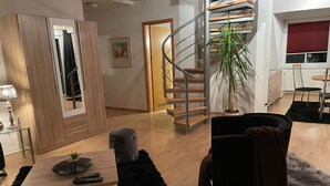 A modern living room with a wooden floor, a black sofa, a small table with a plant, a grey ottoman, and a spiral staircase.