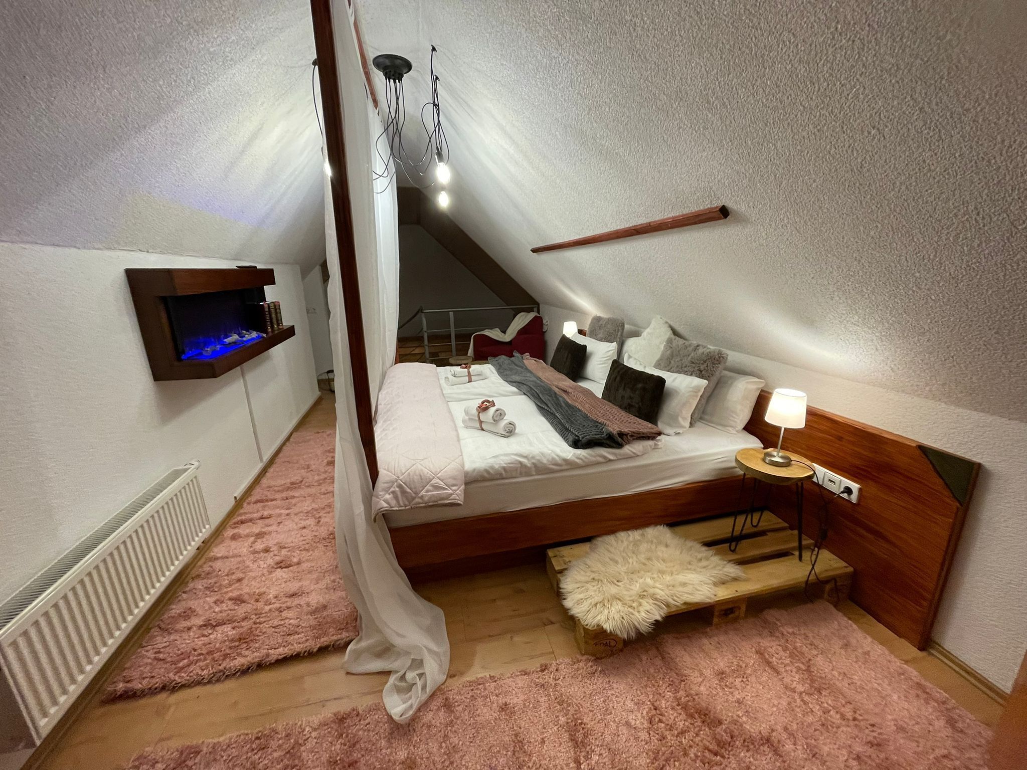 A bedroom with a sloped ceiling, a bed with pillows and a blanket, a bedside table with a lamp, a small shelf with books, and a radiator.