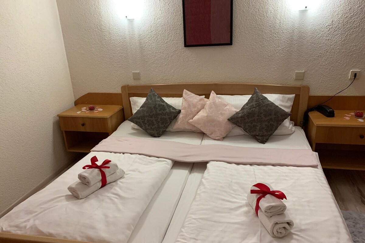 A neatly made double bed with white linens and red bows, two nightstands with lamps, and a wall-mounted light fixture.