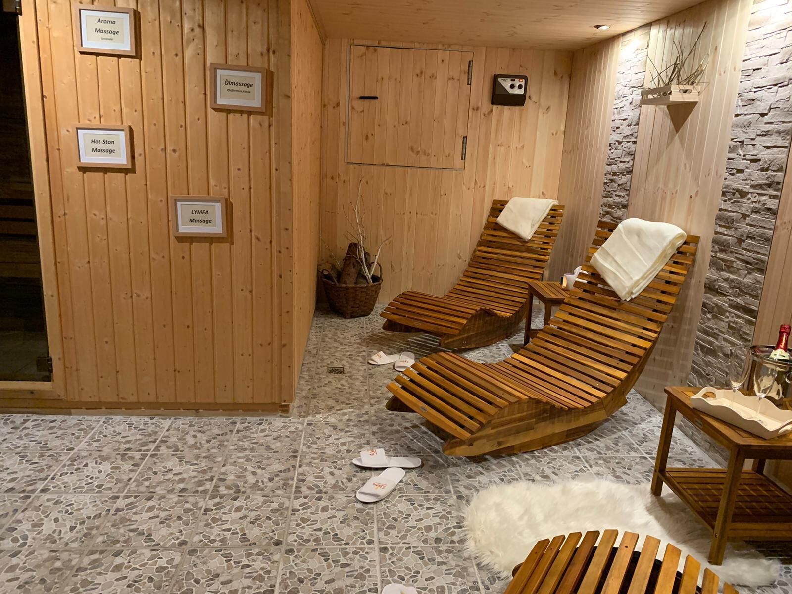 Un spa con paredes de madera, tres tumbonas de madera, suelo de baldosas de piedra y carteles que indican distintos servicios de masaje.