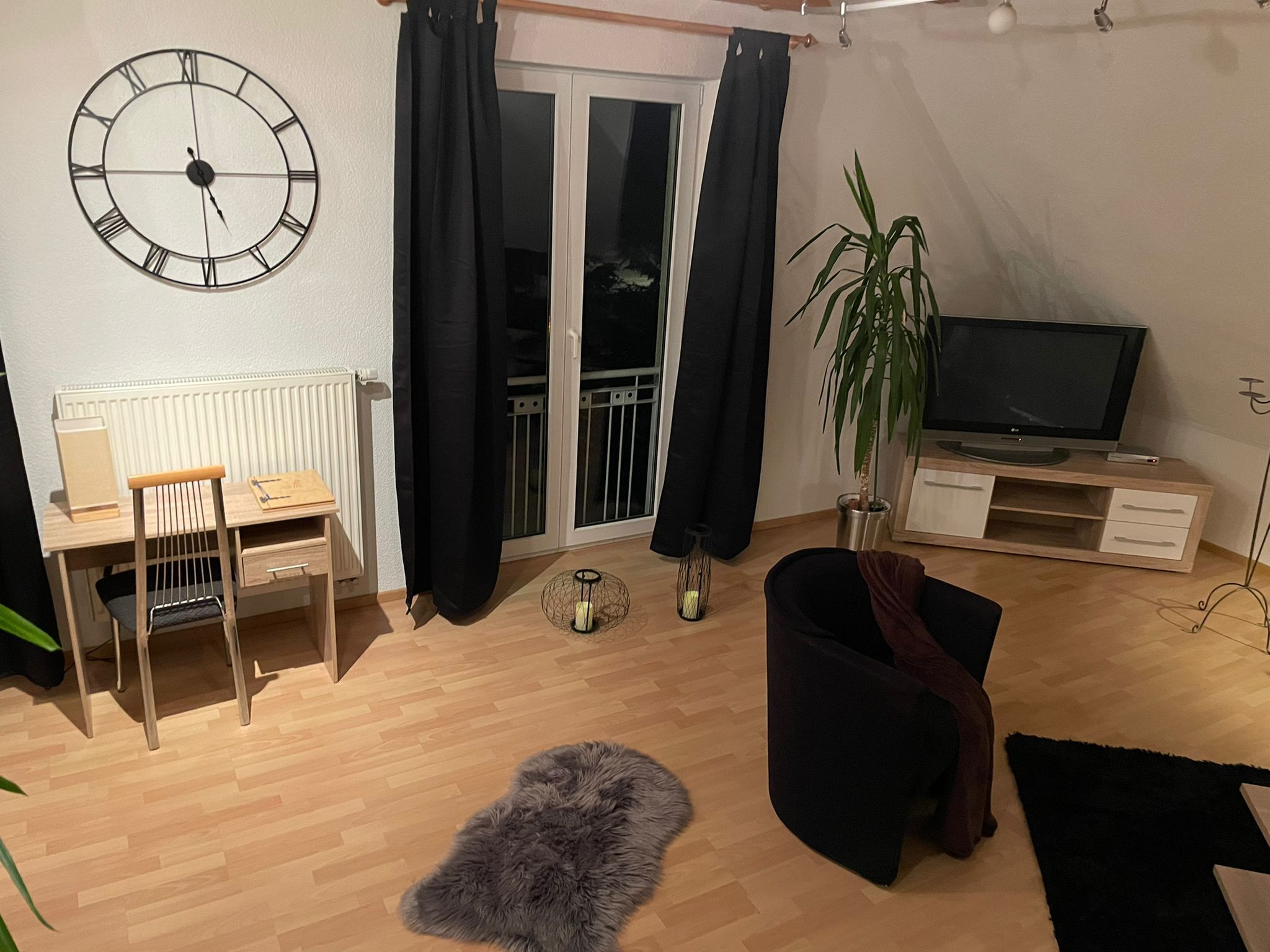 A modern living room with a wooden desk, a black sofa, a TV on a wooden stand, a large clock on the wall, and a sliding glass door.
