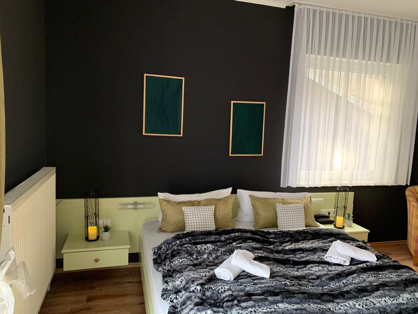 A bedroom with a bed, two bedside tables, a nightstand, a window with white curtains, and two framed pictures on the wall.