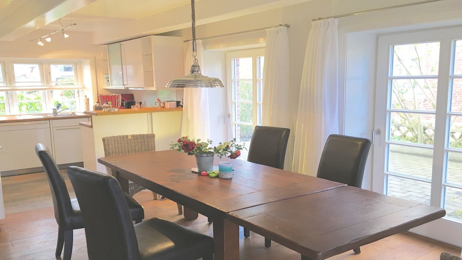 A dining area with a wooden table, black chairs, and a vase with flowers. There is a kitchen area with white cabinets and a hanging light fixture.