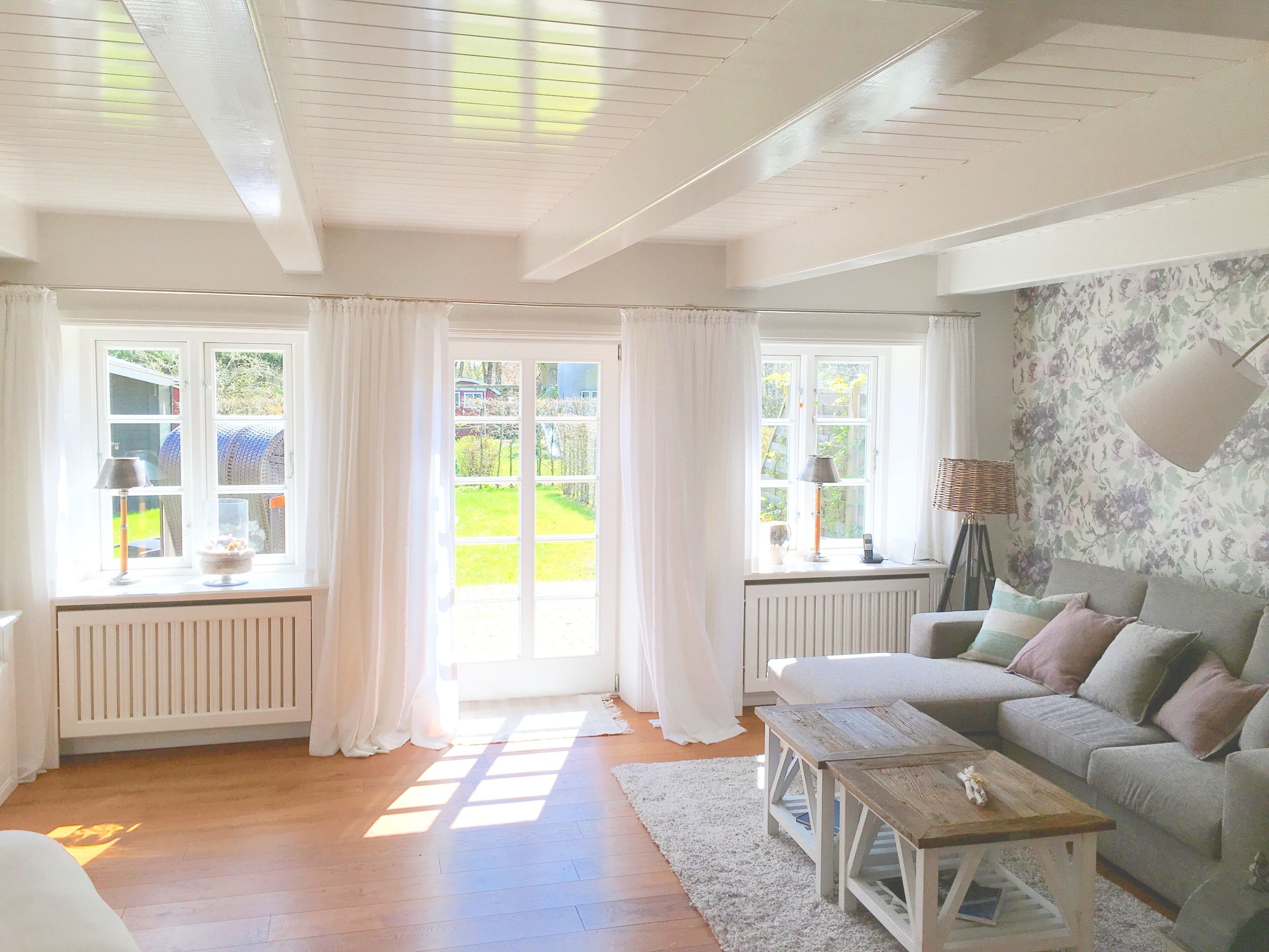 A bright living room with a sofa, a coffee table, and a lamp. There are windows with curtains, a radiator, and a view of greenery outside.