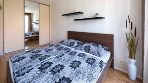 Un dormitorio con una cama grande, un espejo, dos estantes con objetos y un florero con plantas.