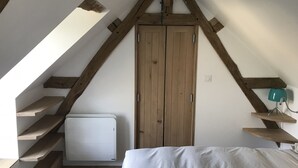 Quarto com teto inclinado, vigas de madeira, radiador branco, cama e uma pequena prateleira com abajur azul-turquesa.