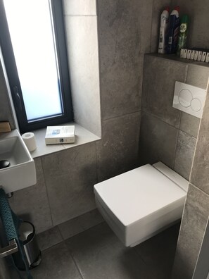 Un baño pequeño con inodoro, lavamanos y ventana. Hay artículos de aseo en el estante sobre el inodoro.