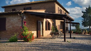 A rustic brick house with a covered patio, a gravel driveway, and a garden with red flowers. - San Lorenzo Agriturismo (Buonconvento)