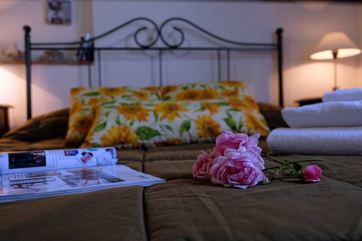 Una cama bien tendida con un cojín floral, una rosa rosa y una revista sobre un edredón marrón.