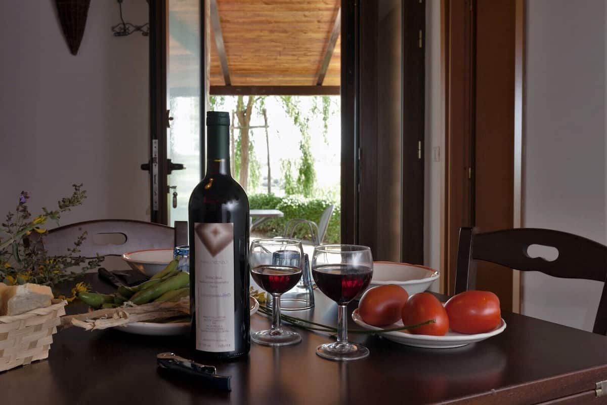 Una mesa con una botella de vino, dos copas de vino tinto, un plato de tomates y una cesta de verduras verdes.