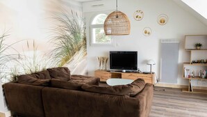 A modern living room with a brown sectional sofa, a flat-screen TV on a wooden console, a wicker pendant light, and a large window with a view of greenery. - Magnificent apartment - Beautiful sea view - Wifi included (Quiberon)