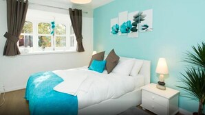 A bedroom with a bed, a nightstand, a lamp, a window with curtains, and a wall with artwork. - Northwick Park Apartment (Harrow)