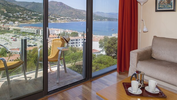 A room with a large window offering a view of a coastal town, a sofa, a coffee table with cups, and wicker chairs. - TORRE GRAND LUXE APARTMENT (El Port de la Selva)