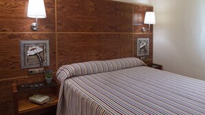 A hotel room with a wooden headboard, two wall-mounted lamps, a bedside table with a lamp, a framed picture, and a striped bedspread. - TORRE GRAND LUXE APARTMENT (El Port de la Selva)