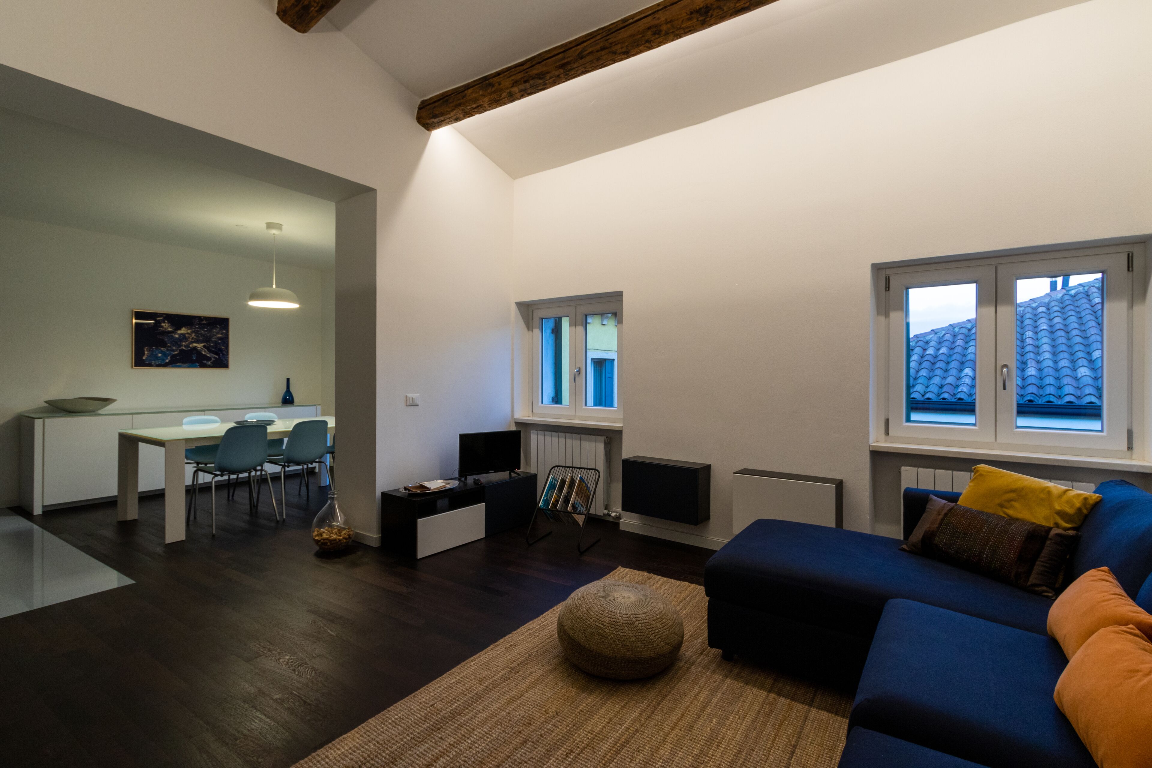 A modern living room with a dark wood floor, a blue sofa, a small rug, and a dining area with a white table and chairs.