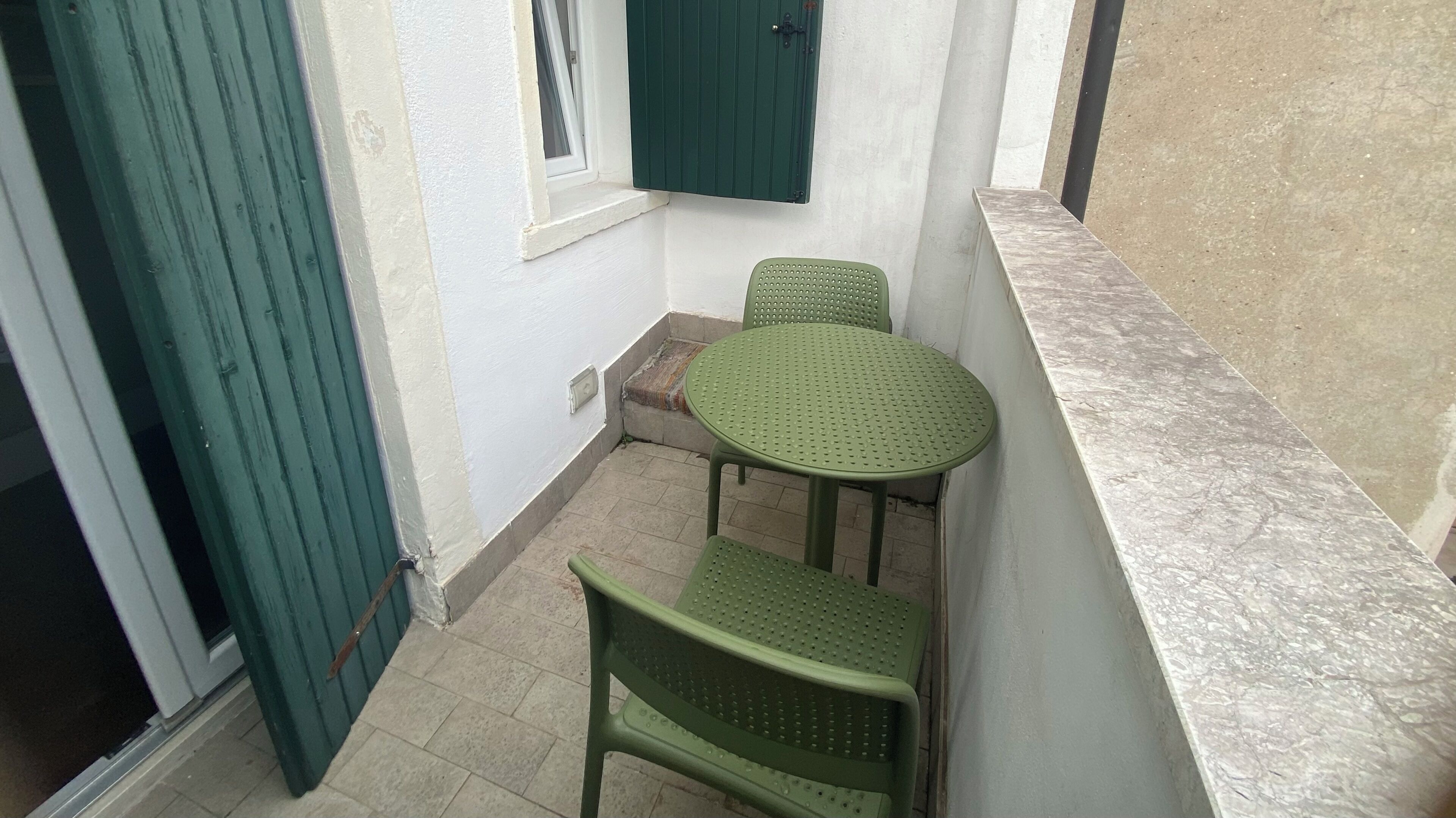 Ein kleiner Außenbereich mit einer grünen Tür, einem Fenster mit grünen Rollläden, einem runden Tisch und zwei grünen Stühlen.