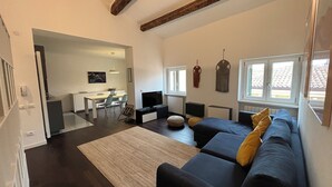 A modern living room with a dark-colored sofa, a small rug, and a dining area with a table and chairs. - Arela (Verona)