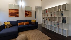 A modern living room with a dark blue sectional sofa, two large desert-themed paintings, a white shelving unit with various items, and a woven rug. - Arela (Verona)