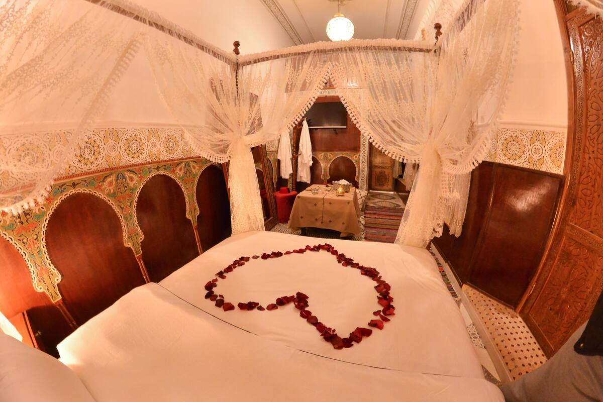 A four-poster bed with a heart-shaped rose arrangement, ornate wooden furniture, and a decorative ceiling.
