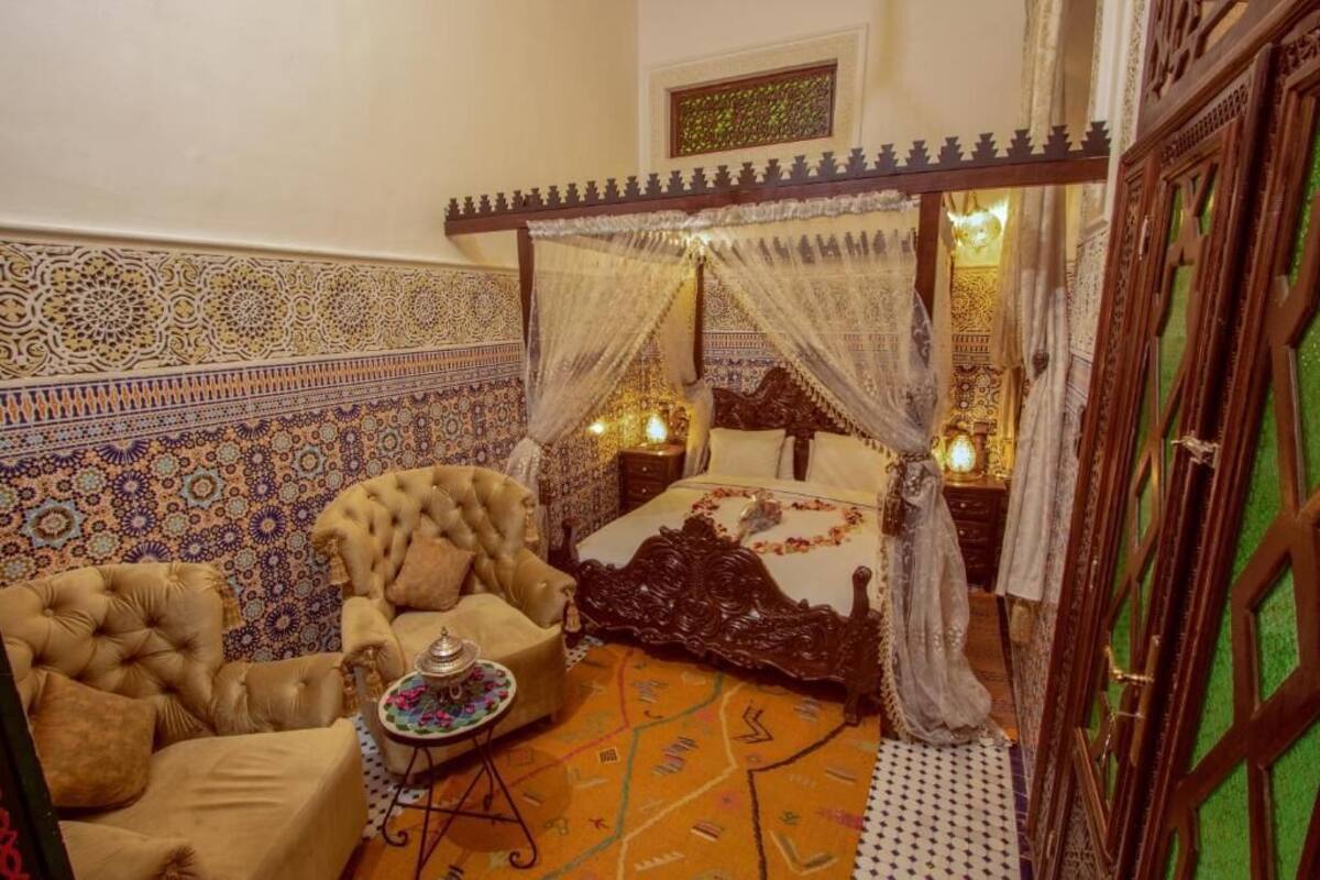 A traditional Moroccan room with a four-poster bed, ornate wall tiles, and a plush armchair.
